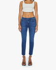 A person stands facing forward in a beige crop top and Mother’s Mid Rise Dazzler Ankle Fray blue jeans, paired with black heeled sandals, against a plain white background.