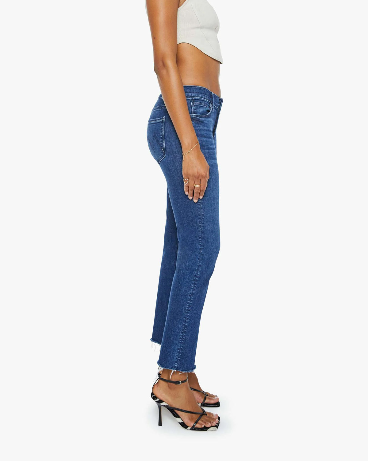 A person wearing a white sleeveless top, Mother Mid Rise Dazzler Ankle Fray jeans, and black strappy high-heeled sandals stands sideways against a plain white background.