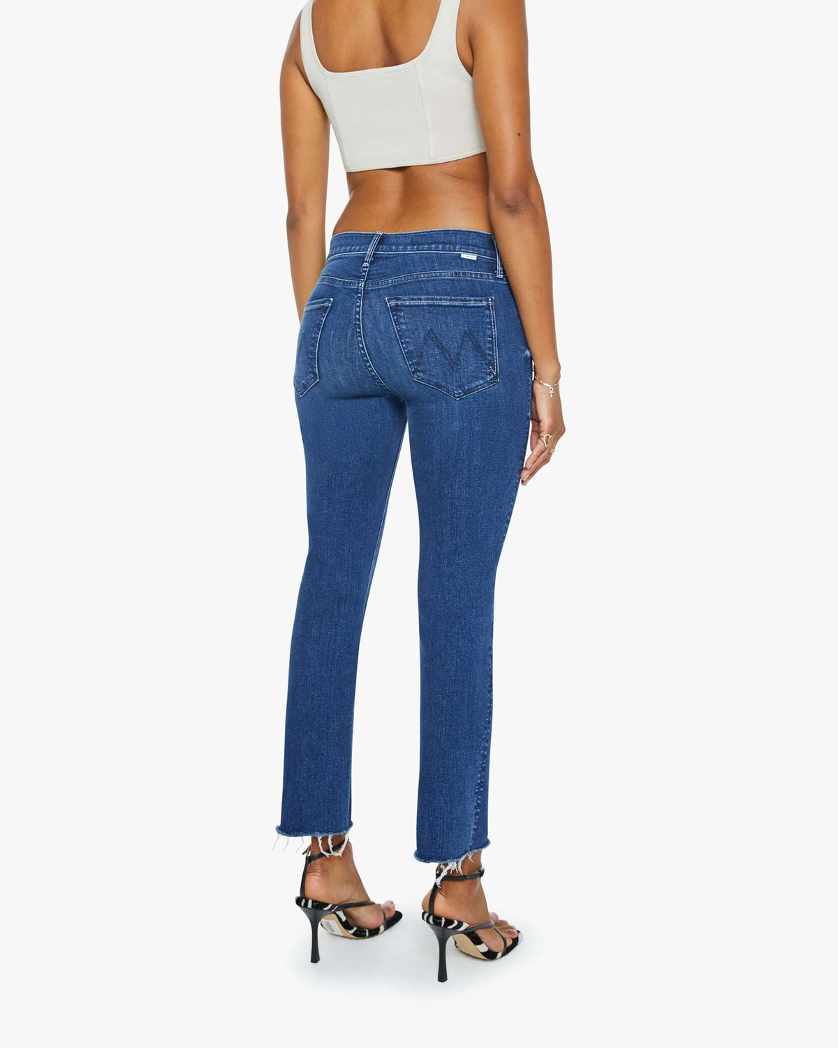 A person wearing a beige crop top, black high-heeled sandals, and Mother’s Mid Rise Dazzler Ankle Fray jeans is standing facing away, highlighting the back details of the jeans.