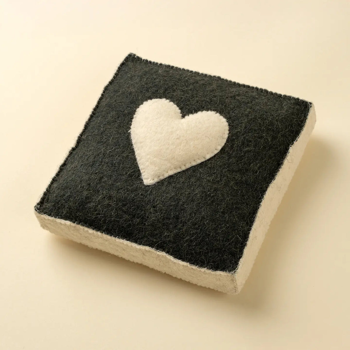 The Indaba Cream Heart Wall Plaque is a square handmade felt decor piece with black sides, a white bottom, and a white heart in the center—displayed on light beige, perfect as unique heart wall art.