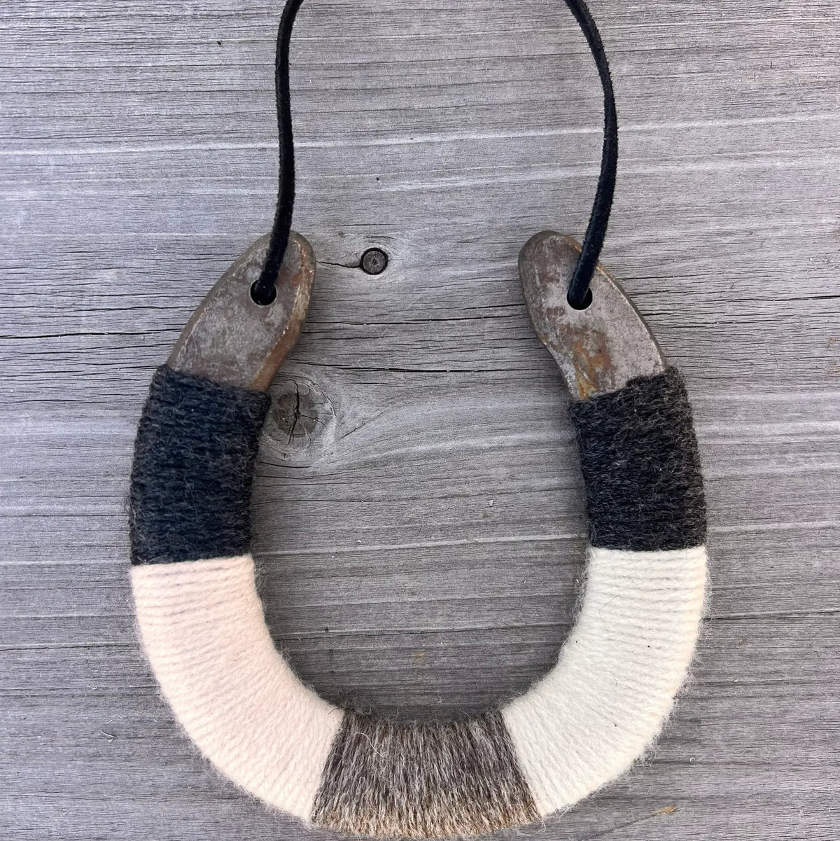 The Lucky Horseshoe by Faire is an upcycled horseshoe wrapped in black, gray, and white yarn, hanging by a black cord against a rustic wooden backdrop—a unique personalized gift.