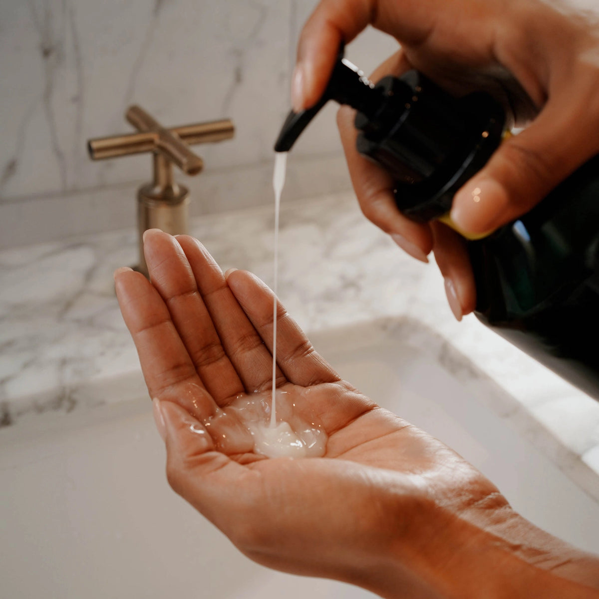 A person dispenses Faire Hand Wash Charlevoix Boreal, a microbiome-friendly liquid soap, from a pump bottle onto their palm over a white sink with a marble countertop and brass faucet in the background.