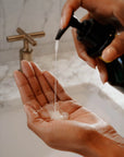A person dispenses Faire Hand Wash Charlevoix Boreal, a microbiome-friendly liquid soap, from a pump bottle onto their palm over a white sink with a marble countertop and brass faucet in the background.