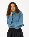 A woman with long curly hair models the Kerri Rosenthal Denim Jacket, featuring heart elbow patches, paired with a black skirt and standing against a plain white background.