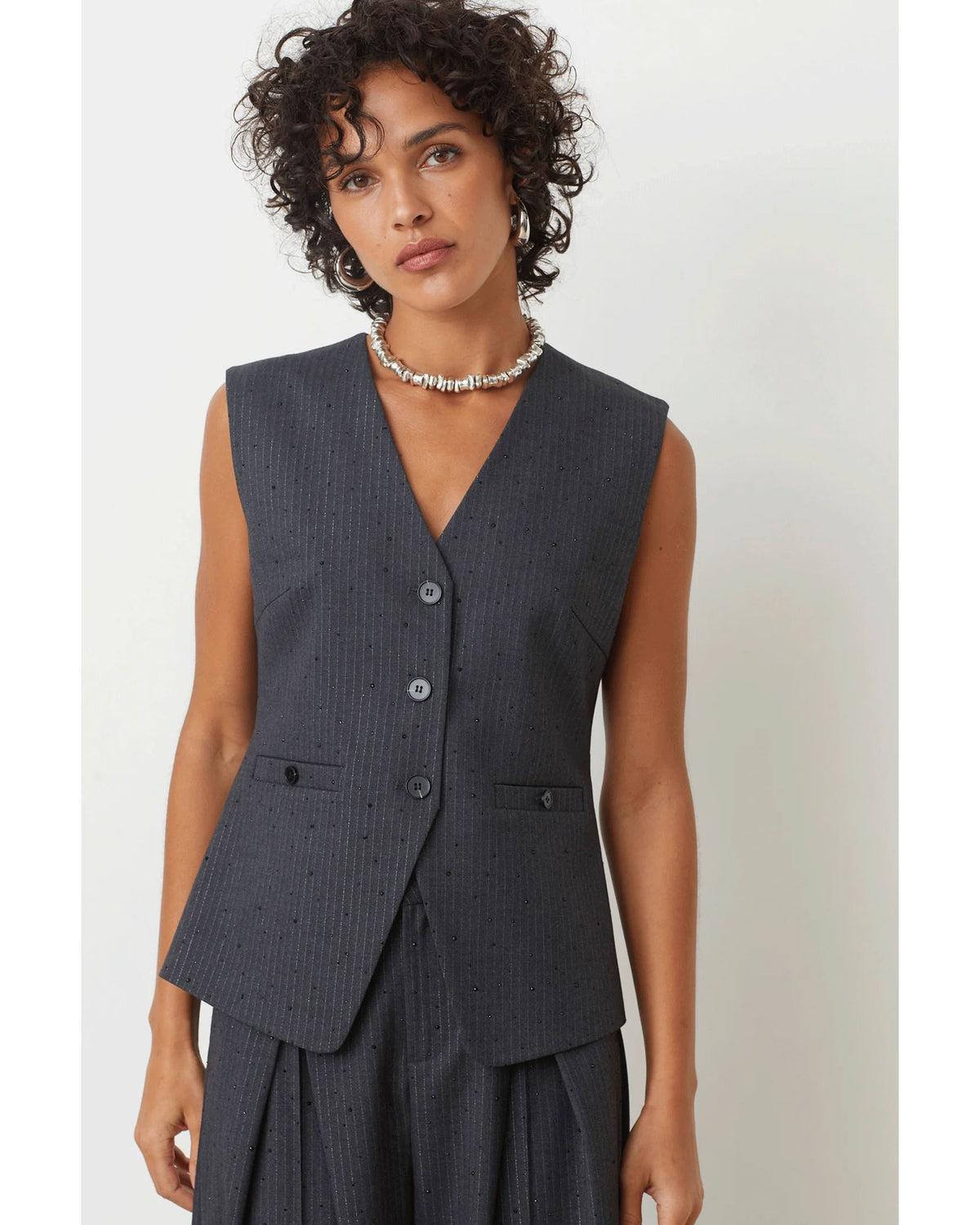 A woman with short curly hair wears the SUNCOO Derek Vest—a sleeveless, dark gray pinstripe piece woven with metallic thread—paired with matching pants and a beaded necklace, standing against a plain white background.