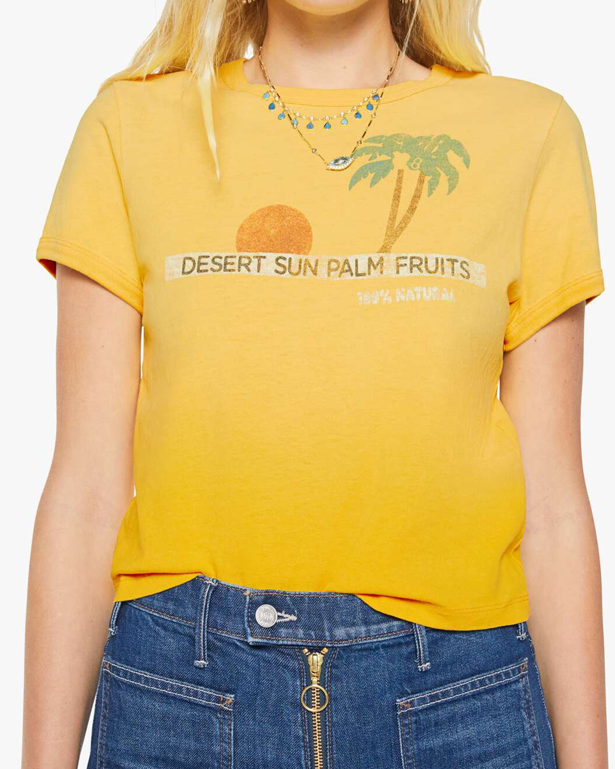 A woman wears the Mother "The Itty Bitty Ringer" tee, a sun-faded yellow shrunken crewneck with a sun and palm trees graphic and "DESERT SUN PALM FRUITS 100% NATURAL" text, paired with blue jeans with visible button and zipper.