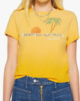A woman wears the Mother "The Itty Bitty Ringer" tee, a sun-faded yellow shrunken crewneck with a sun and palm trees graphic and "DESERT SUN PALM FRUITS 100% NATURAL" text, paired with blue jeans with visible button and zipper.