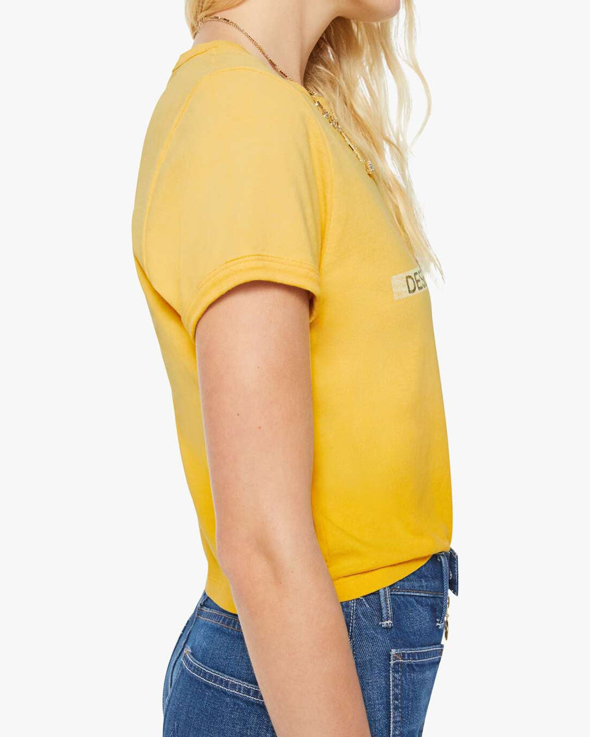 A woman with wavy blonde hair is shown from the side, wearing The Itty Bitty Ringer crop tee by Mother in yellow and blue jeans against a plain white background.