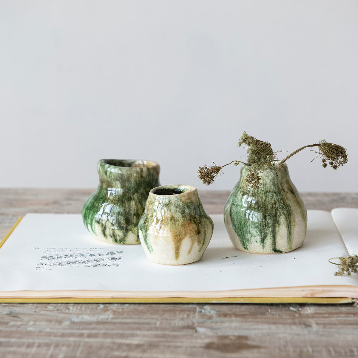 Three small Crackle Glaze Vases by Creative Co-op, in green and beige with a reactive crackle finish, sit on an open book atop wood. One holds dried flowers. The plain, neutral background highlights the organic shapes.