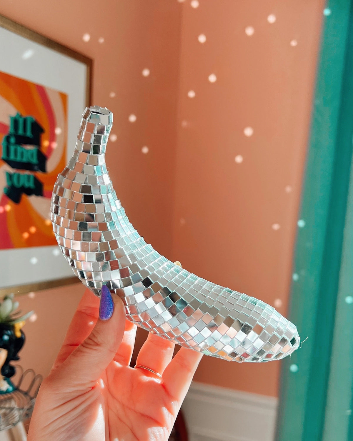 A hand with purple nail polish holds the Disco Banana by Faire—a banana-shaped decor piece covered in glass mirror tiles—casting shimmering reflections on a peach-colored wall in the background.