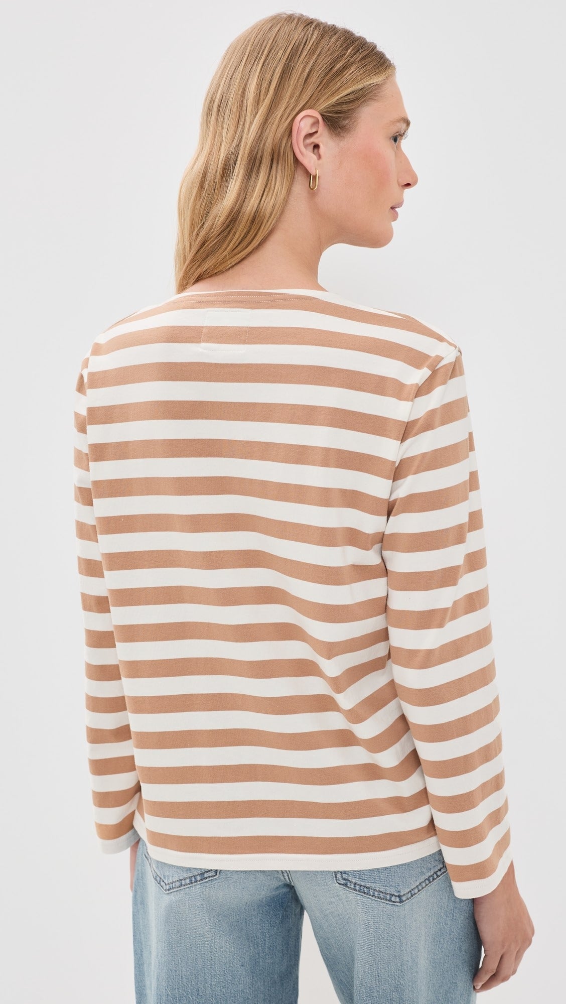 A woman with long blonde hair and gold hoop earrings wears the Denimist MILLS POND STRIPED T in beige and white stretch jersey with light blue jeans, standing with her back to the camera.