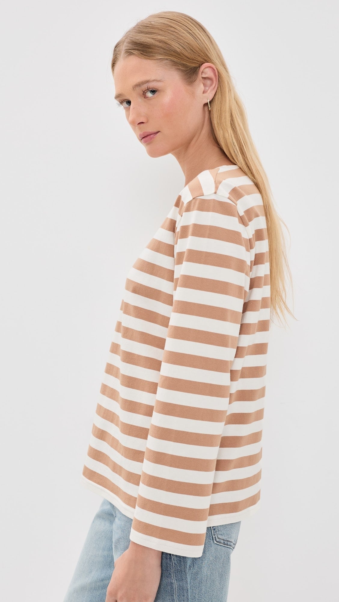 A woman with long blonde hair models the Denimist MILLS POND STRIPED T, a beige and white lightweight jersey tee, paired with light blue jeans. She stands sideways, facing the camera against a plain white background.