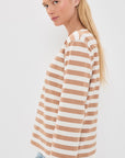 A woman with long blonde hair models the Denimist MILLS POND STRIPED T, a beige and white lightweight jersey tee, paired with light blue jeans. She stands sideways, facing the camera against a plain white background.