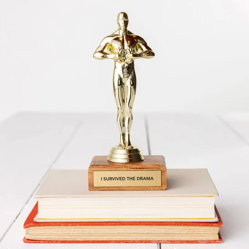 A JE Trophy resembling an Oscar statuette stands on top of stacked books, with a plaque reading "I SURVIVED THE DRAMA." The background is a plain white surface in Arizona-style.