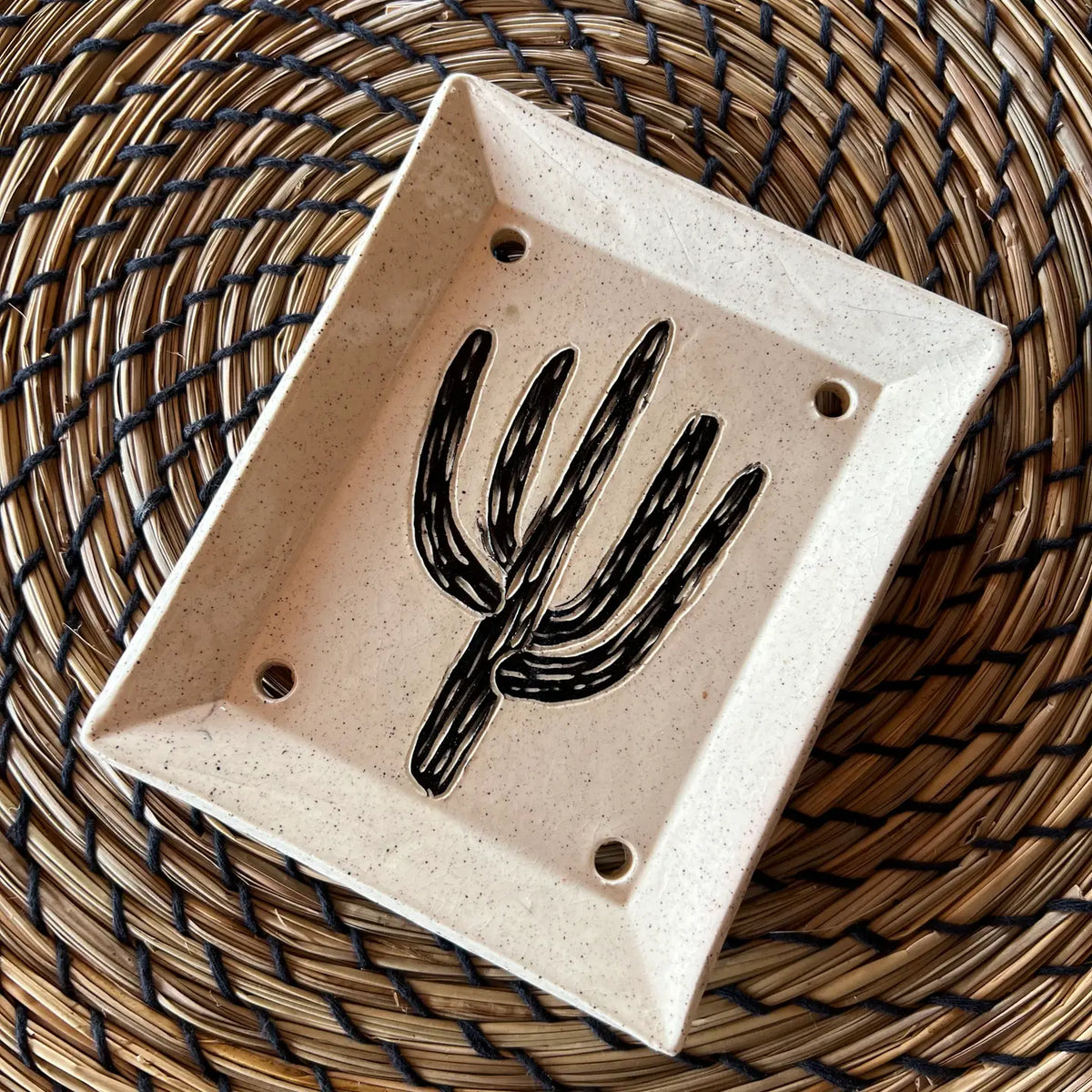 The Saguaro Soap Dish, a handmade piece with a black sgraffito cactus design and four drainage holes, rests on a woven spiral-patterned placemat.