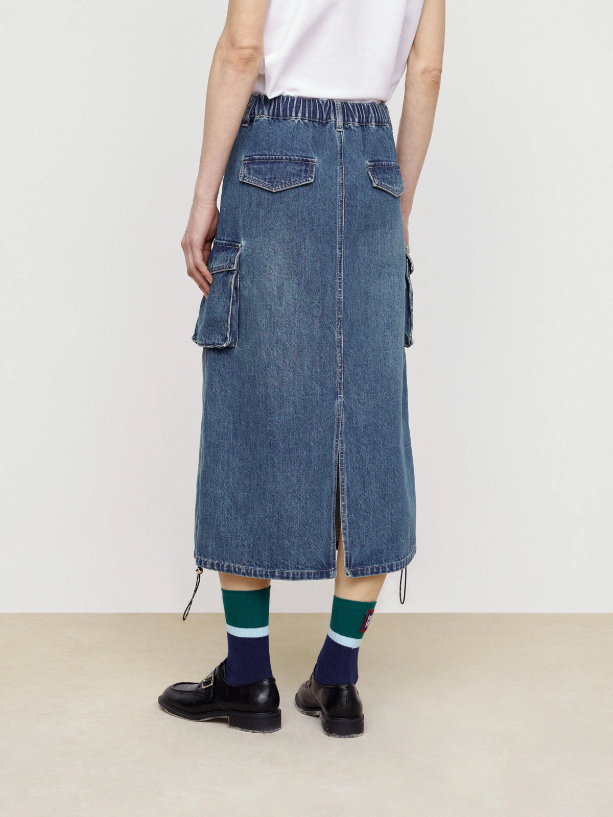 A person in street style wears a white shirt and the Faire Pocket Denim Half Skirt, featuring workwear pockets, back slit, and drawstring hem, styled with dark shoes and blue, green, and white socks. Their back faces the camera.