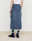 A person in street style wears a white shirt and the Faire Pocket Denim Half Skirt, featuring workwear pockets, back slit, and drawstring hem, styled with dark shoes and blue, green, and white socks. Their back faces the camera.