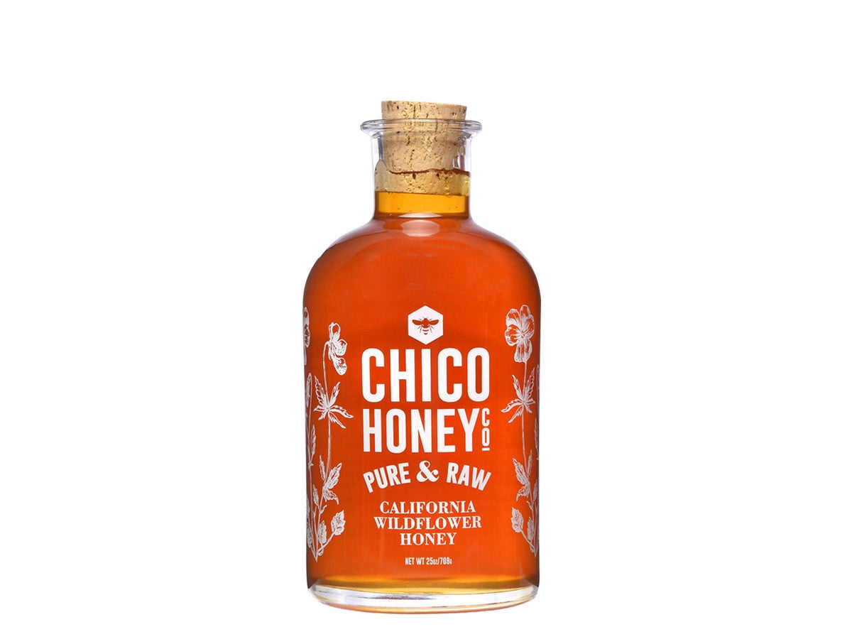 A clear glass bottle of Olivarez Chico Honey by Faire with a cork stopper, featuring white floral designs and text on the front label.