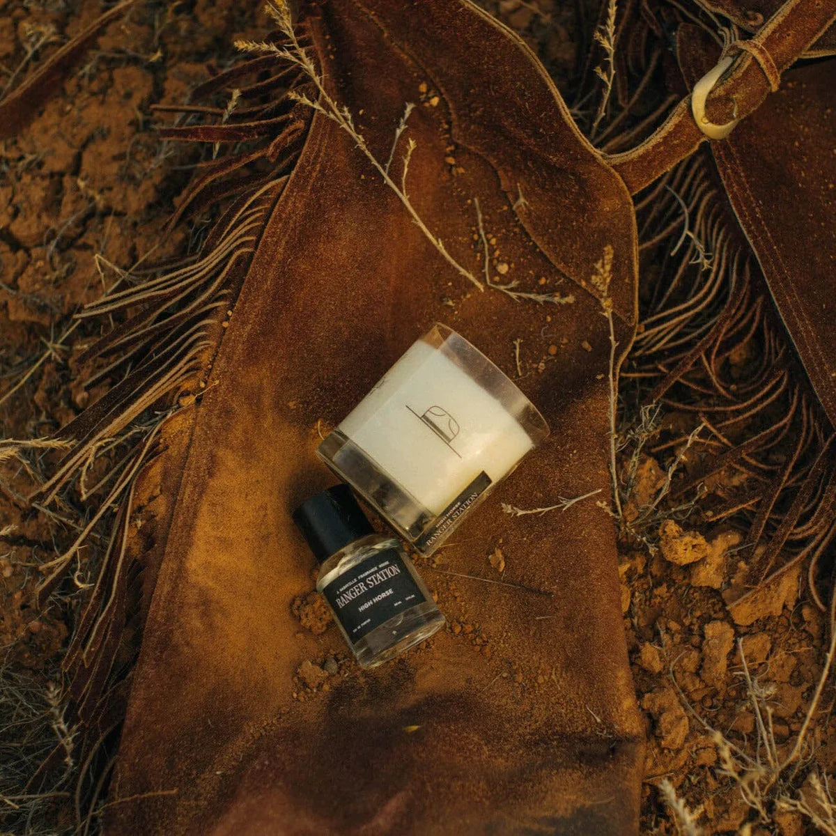 High Horse candle and fragrance bottle displayed on brown leather fringe chaps over rocky ground.