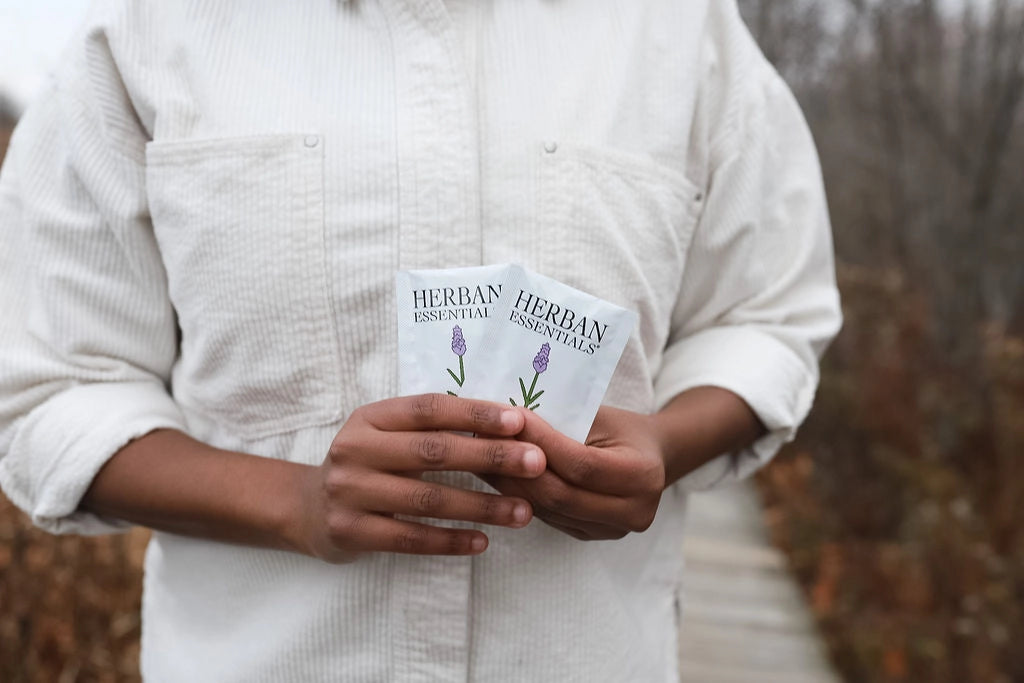 A person in a white shirt holds two packets of Faire Lavender Towelettes – 20 Individual Essential Oil Wipes, each with a lavender illustration, while standing outdoors on a wooden path amid autumn foliage.