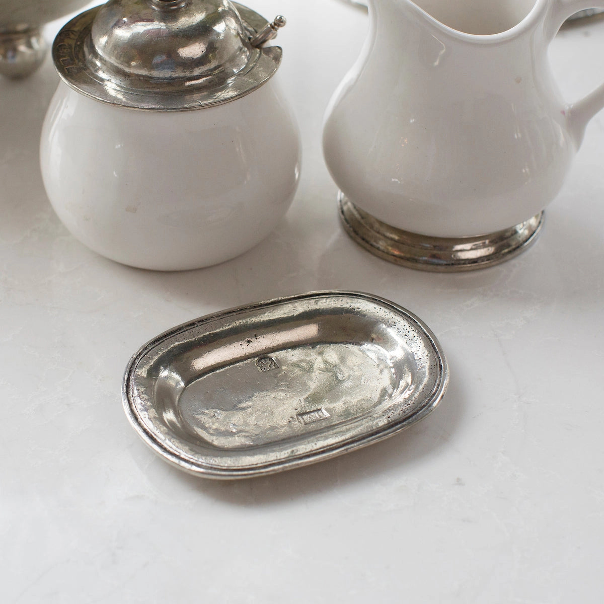The Faire Vintage Bedside Tray—a small, rectangular silver dish crafted from Italian pewter—rests on a white surface beside a white ceramic pitcher and a lidded container with silver accents, evoking the charm of an elegant jewelry organizer or classic bedside accessory.