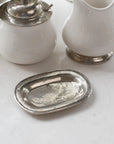 The Faire Vintage Bedside Tray—a small, rectangular silver dish crafted from Italian pewter—rests on a white surface beside a white ceramic pitcher and a lidded container with silver accents, evoking the charm of an elegant jewelry organizer or classic bedside accessory.