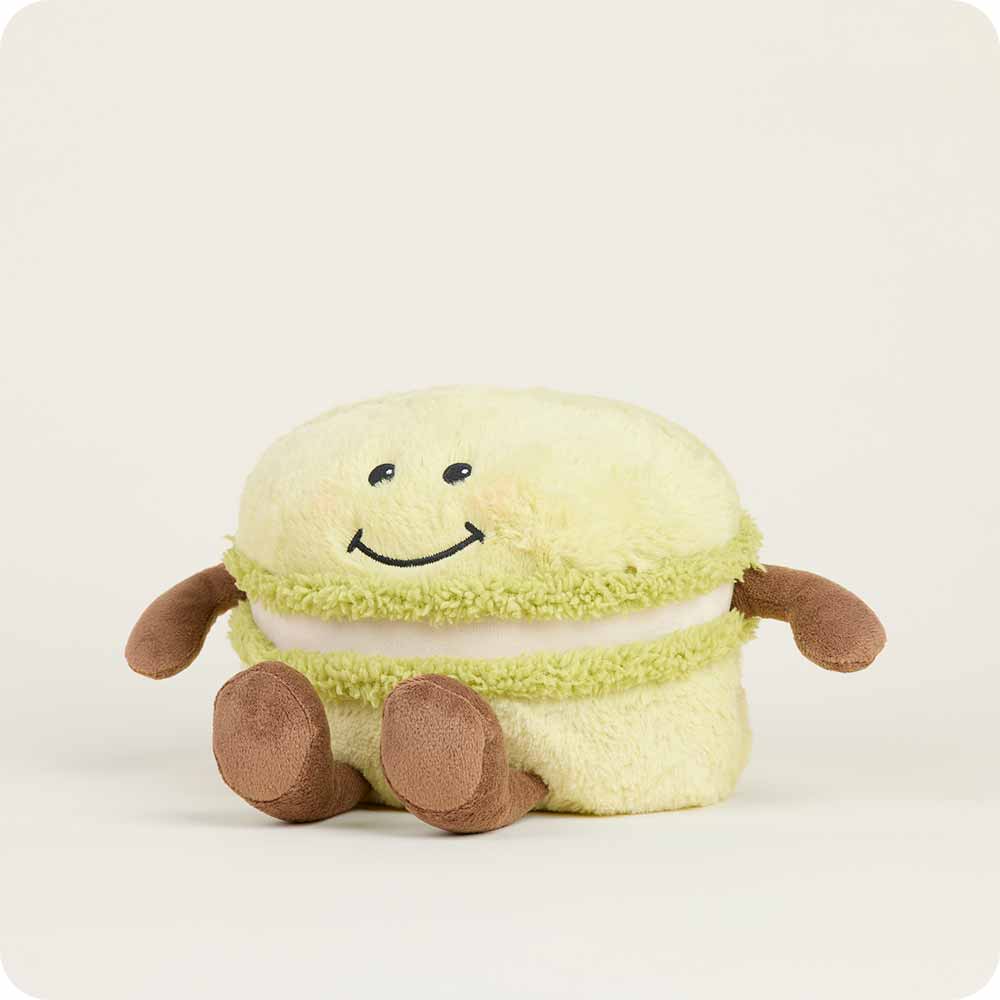 The Faire Green Macaroon Warmie is a microwavable plush toy shaped like a light green macaron with a smiling face, brown arms, and legs, offering sensory comfort against a plain light background.