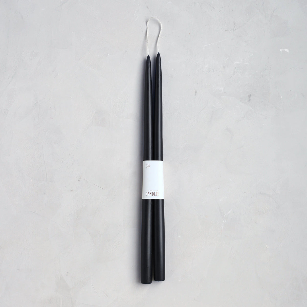 The Floral Society’s 18" Taper Candle - Pair is wrapped in a white paper label, set against a light gray textured background—perfect for adding an elegant touch to any home décor.