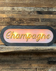 The Faire Lighted Sign features the word "Champagne" in cursive neon, glowing against a rustic wooden wall—ideal as personalized wall decor or a custom illuminated sign for your space.
