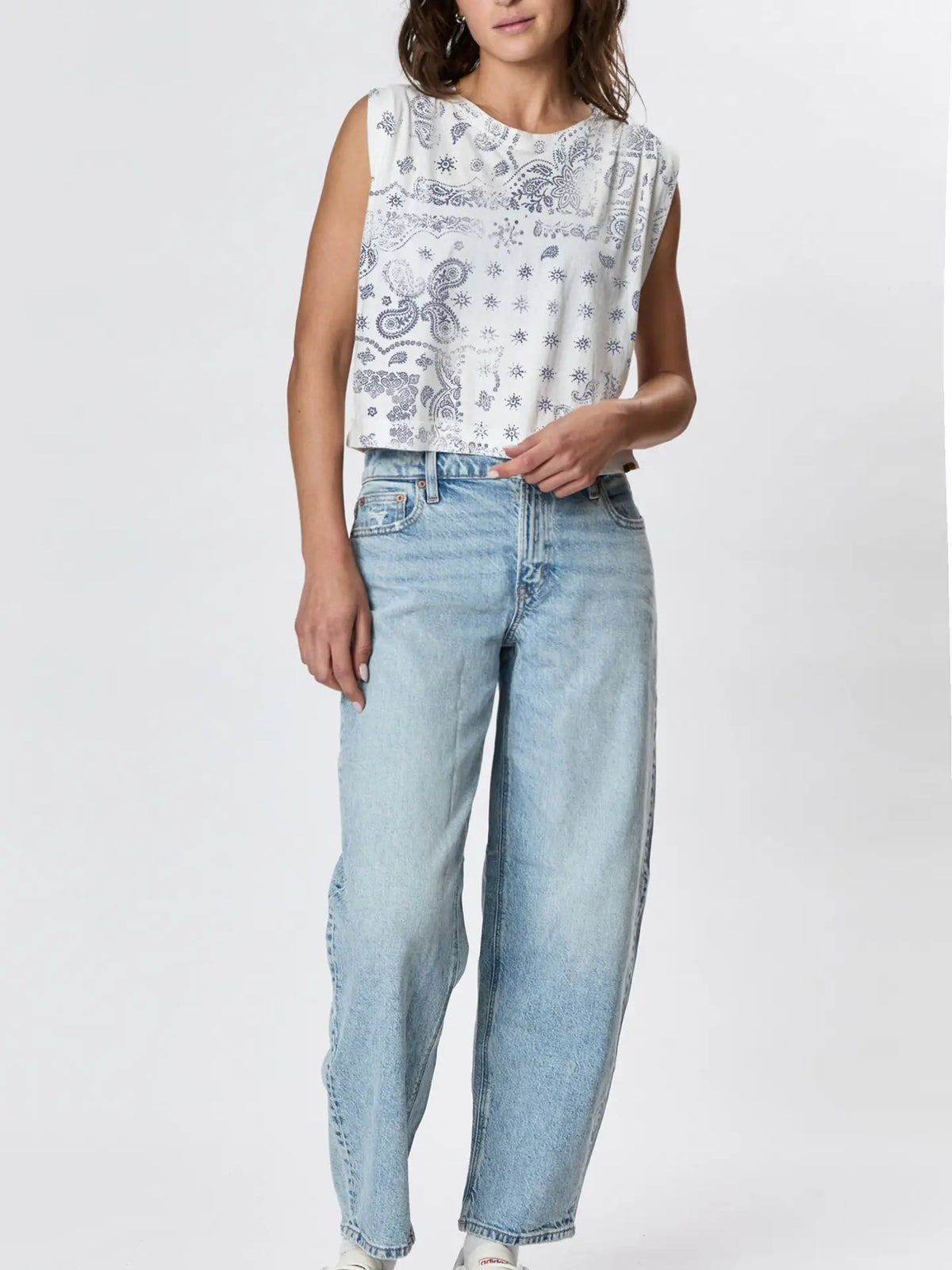 A woman wears the Faire Hemp Jackie Pleat Shell Bandana, a sleeveless white hand-printed shirt with blue paisley and star patterns, paired with light blue high-waisted relaxed-fit jeans, standing against a plain white background.