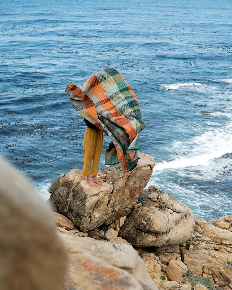 A person stands barefoot on a rocky ocean cliff, wrapped in the Faire Vrou-Vrou blanket with bold textile design blowing in the wind, as vivid blue waves and swirling seaweed move below.