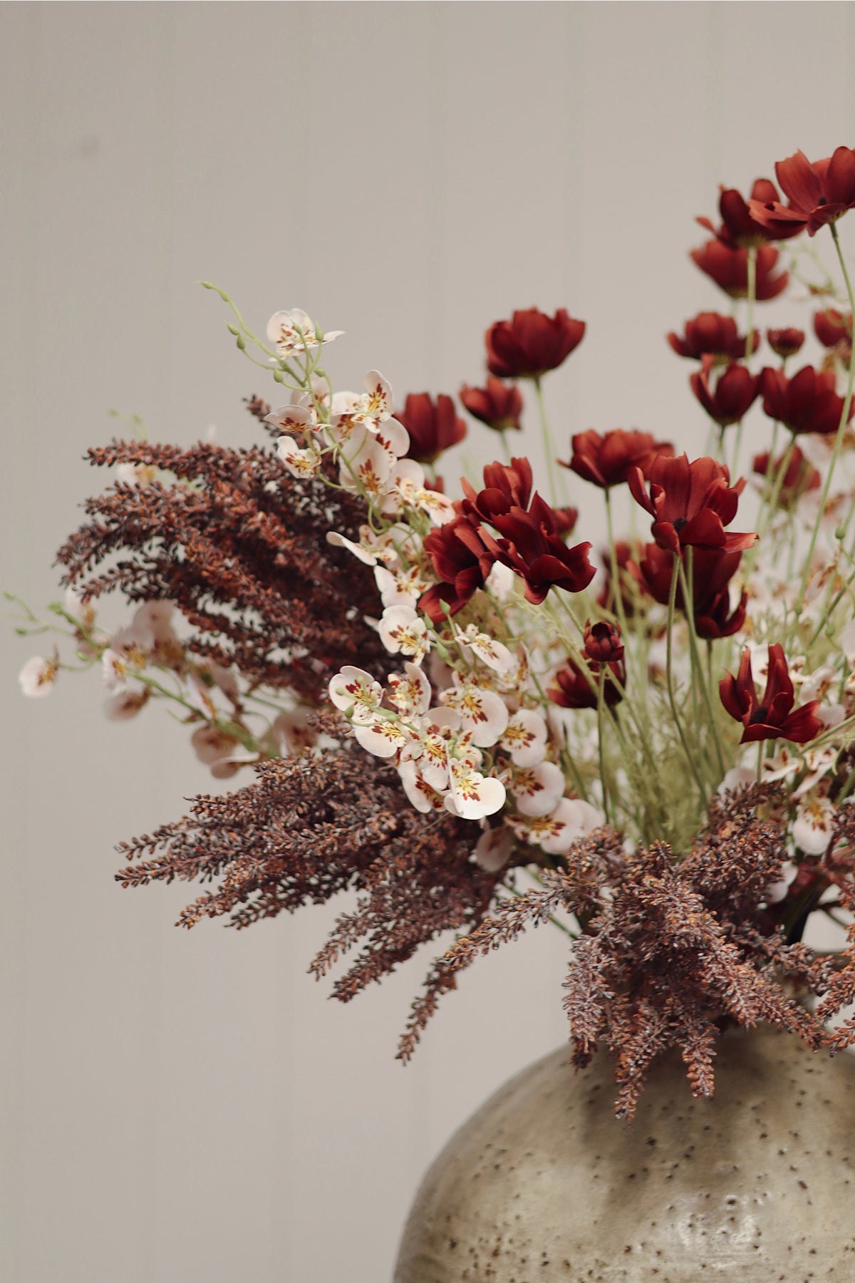 The Faire Calliopsis Spray in rust red features faux flowers with delicate white blossoms and wispy brown foliage, artfully arranged in a ceramic vase against a neutral light background.