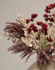 The Faire Calliopsis Spray in rust red features faux flowers with delicate white blossoms and wispy brown foliage, artfully arranged in a ceramic vase against a neutral light background.