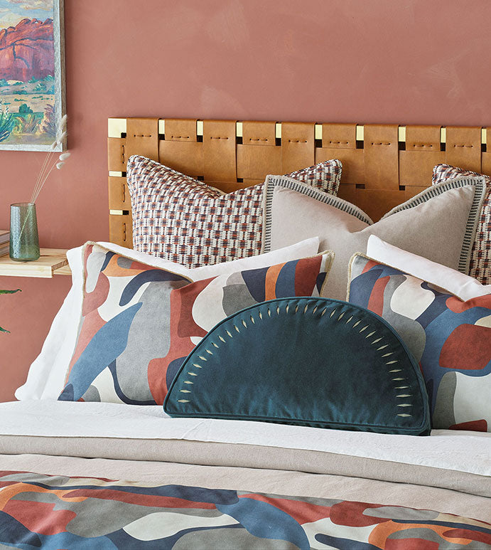 A stylishly decorated bedroom corner in a Scottsdale bungalow featuring a Moab Graphic 27x27" bedspread from Eastern Accents with an artistic headboard against a soft coral wall, adorned with decorative pillows in various patterns and a bedspread with abstract design.