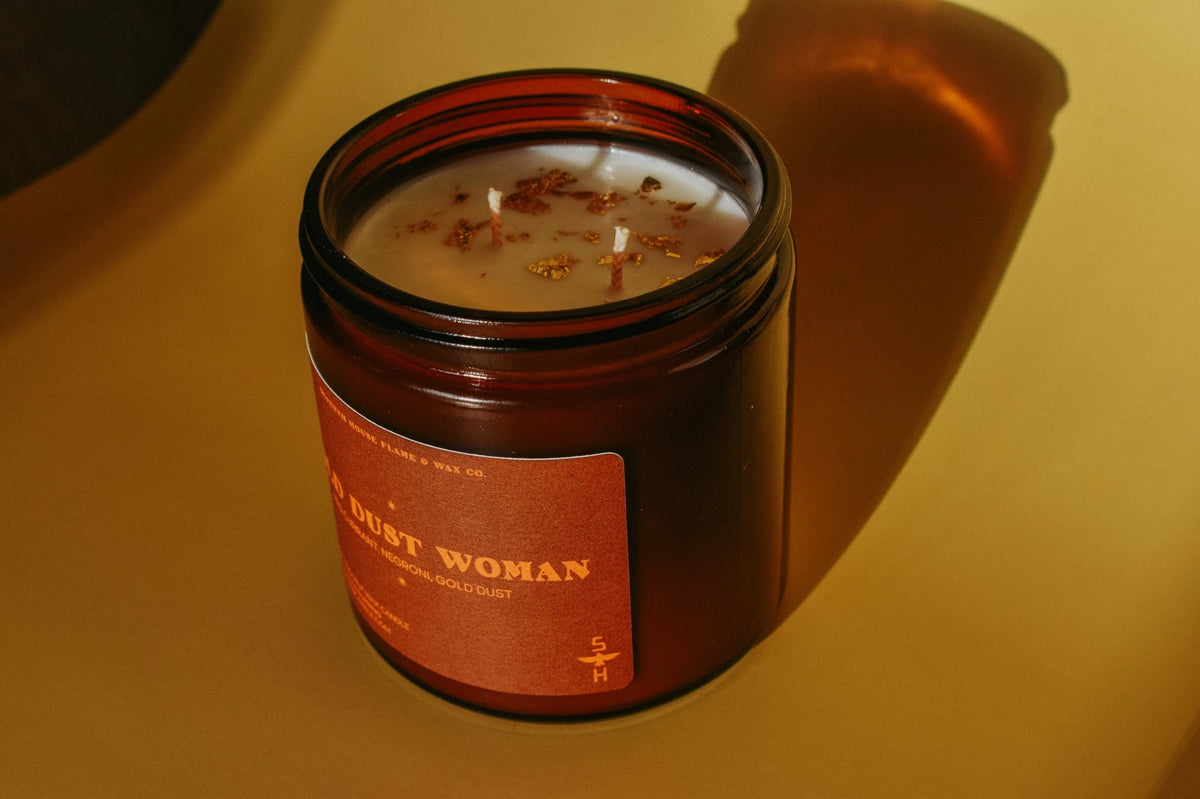 Gold Dust Woman Candle by Faire with gold-flecked wax, casting soft shadows on a yellow tabletop.