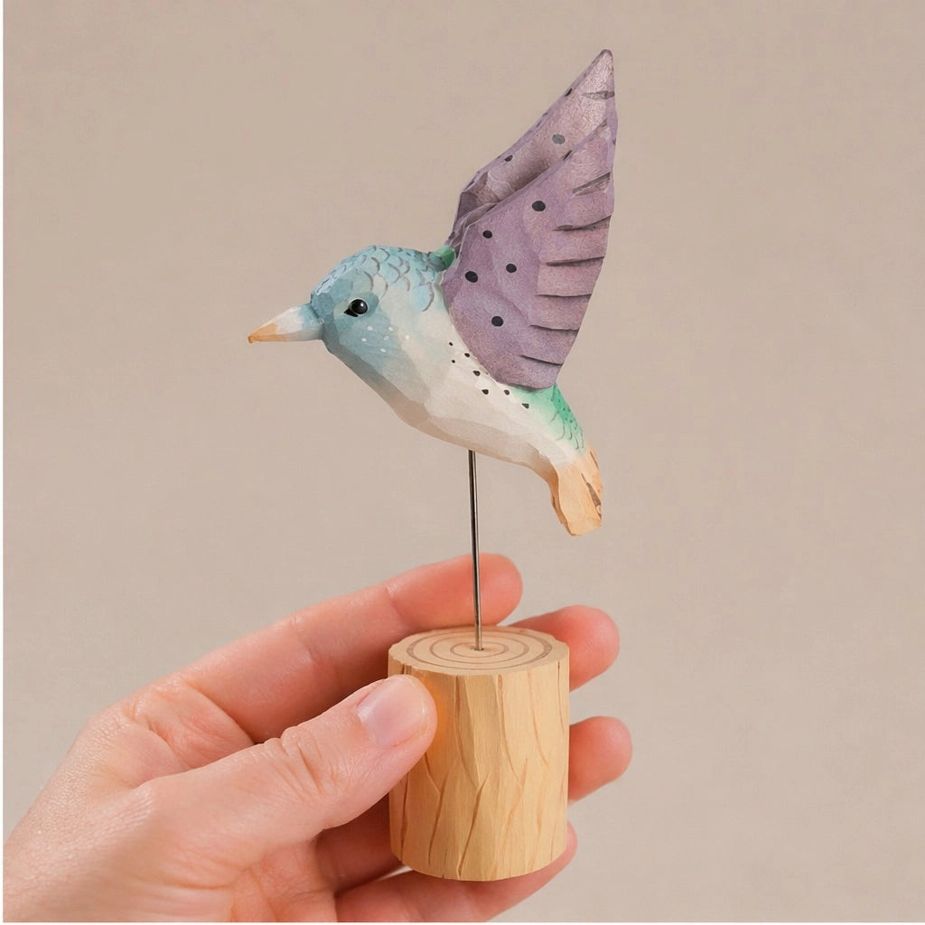 Purple hand-painted carved hummingbird figurine on wood base by Faire, shown held in hand for scale.