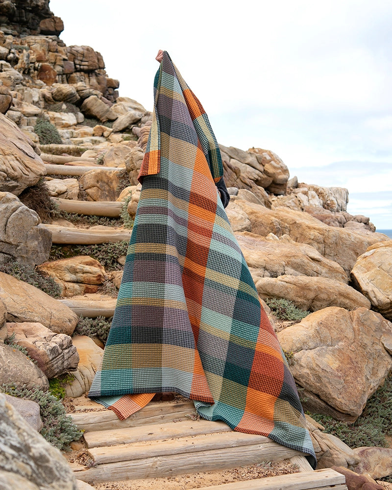 Wrapped in a vibrant Faire Vrou-Vrou blanket featuring bold orange, green, yellow, and gray patterns, a person stands on rocky outdoor steps beneath a partly cloudy sky.