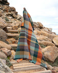 Wrapped in a vibrant Faire Vrou-Vrou blanket featuring bold orange, green, yellow, and gray patterns, a person stands on rocky outdoor steps beneath a partly cloudy sky.