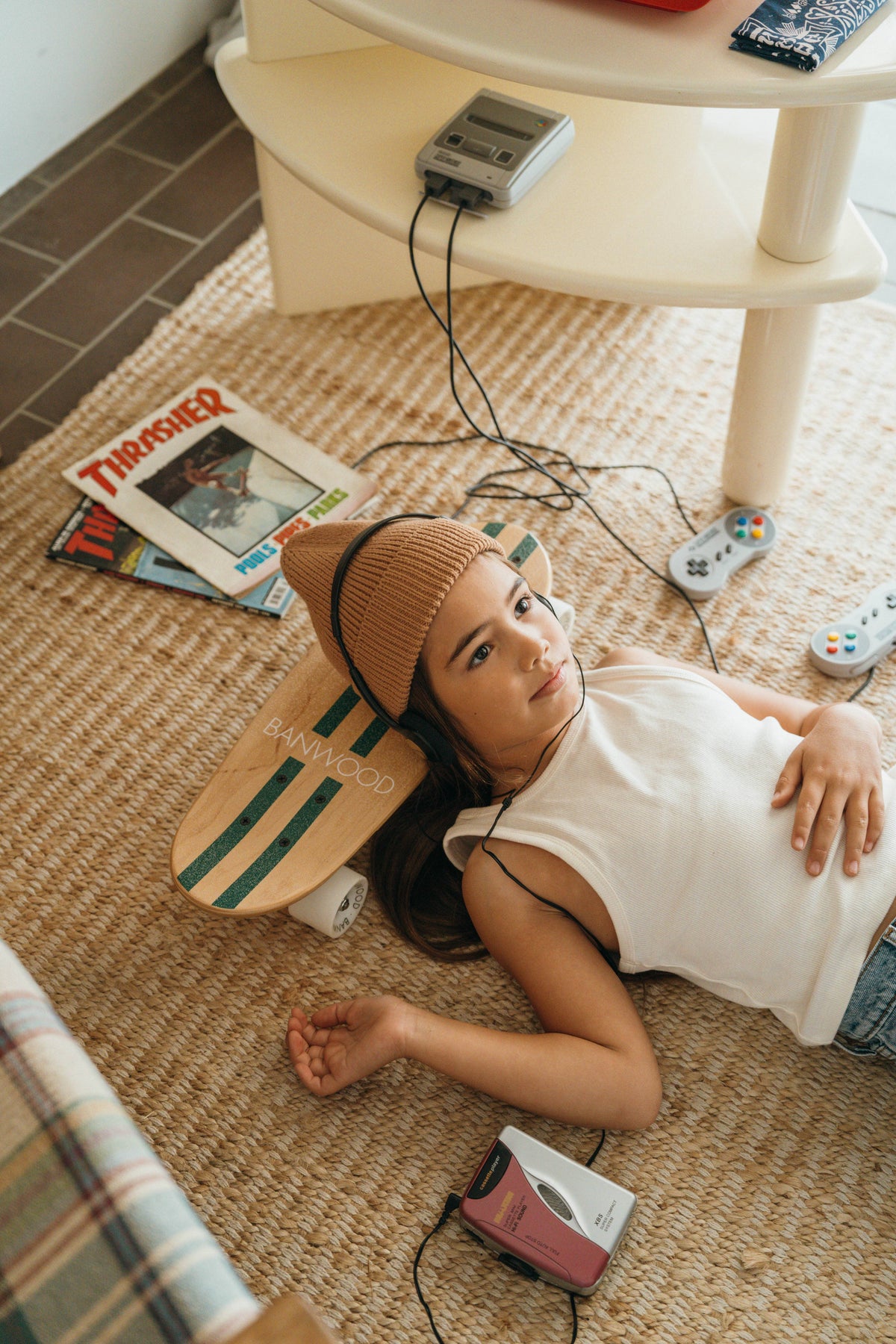 A girl in a beanie and tank top relaxes on a woven rug with headphones, resting her head on the BANWOOD SKATEBOARD - GREEN by Faire. Vintage video game controllers, a cassette player, and skateboard magazines are nearby.