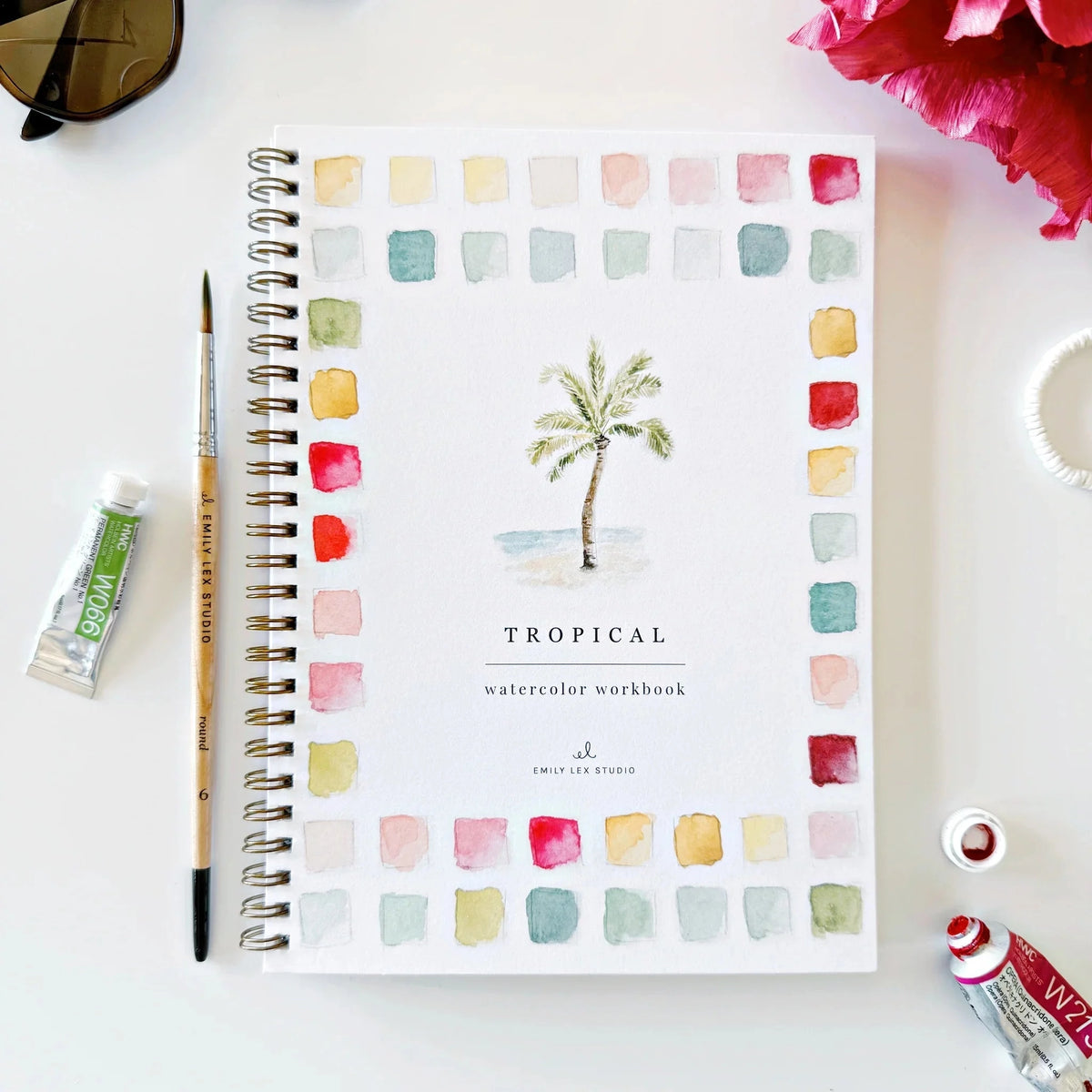 The Faire Watercolor Workbook is a spiral-bound beginner’s guide featuring a palm tree illustration and colorful swatches on the cover, shown with art supplies, a flower, sunglasses, and a hair tie on a white surface.