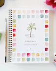 The Faire Watercolor Workbook is a spiral-bound beginner’s guide featuring a palm tree illustration and colorful swatches on the cover, shown with art supplies, a flower, sunglasses, and a hair tie on a white surface.