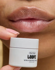 A close-up of lips with a glossy finish shows a small white jar labeled "Faire Dew Cloud Mini Lip Mask" near the mouth, highlighting this ultra-moisturizing, hydrating lip treatment.