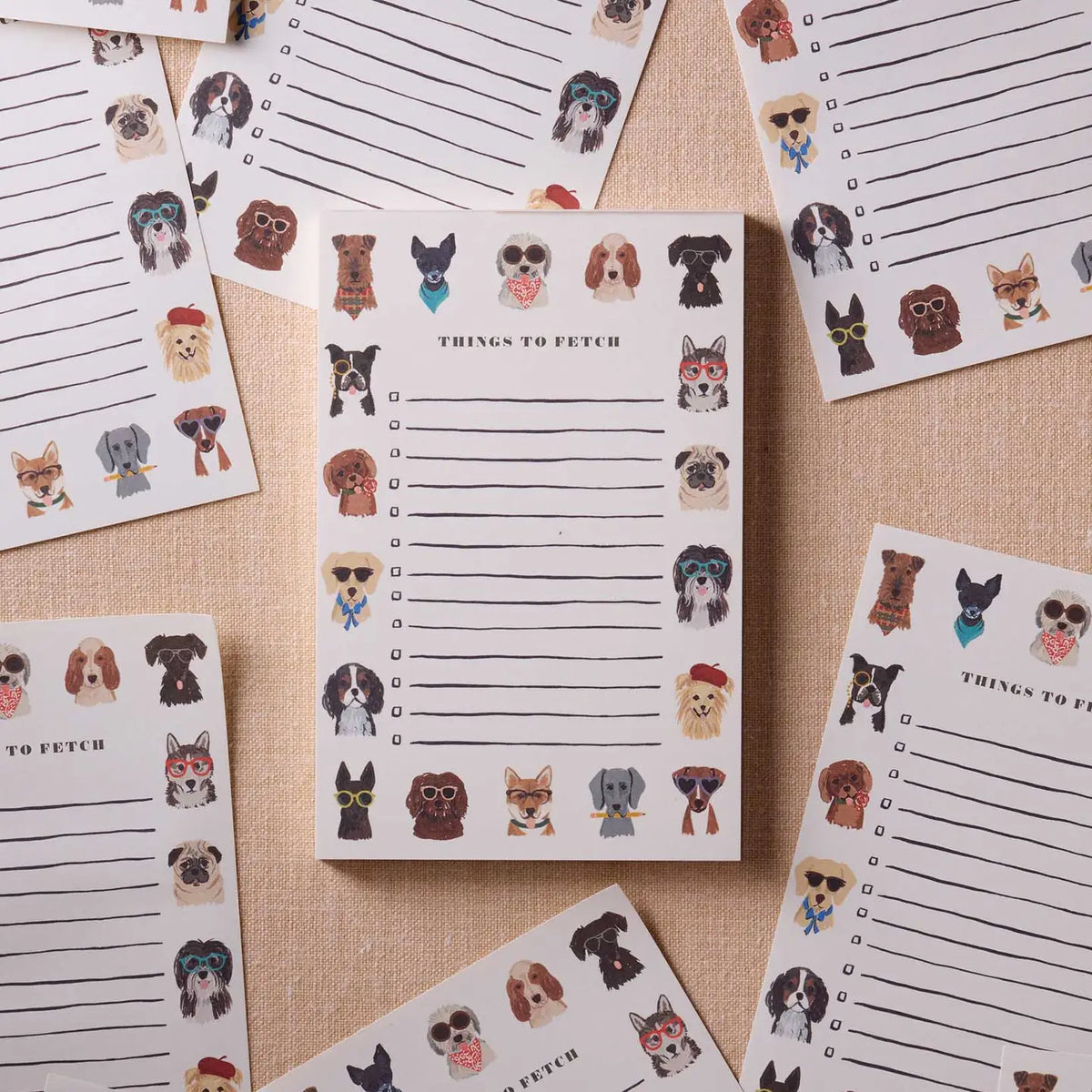 The Dog Days Notepad by Faire features cute dog faces along the top and bottom. Multiple tear-off sheets are shown on a neutral fabric, each perfect for jotting down playful to-do lists.