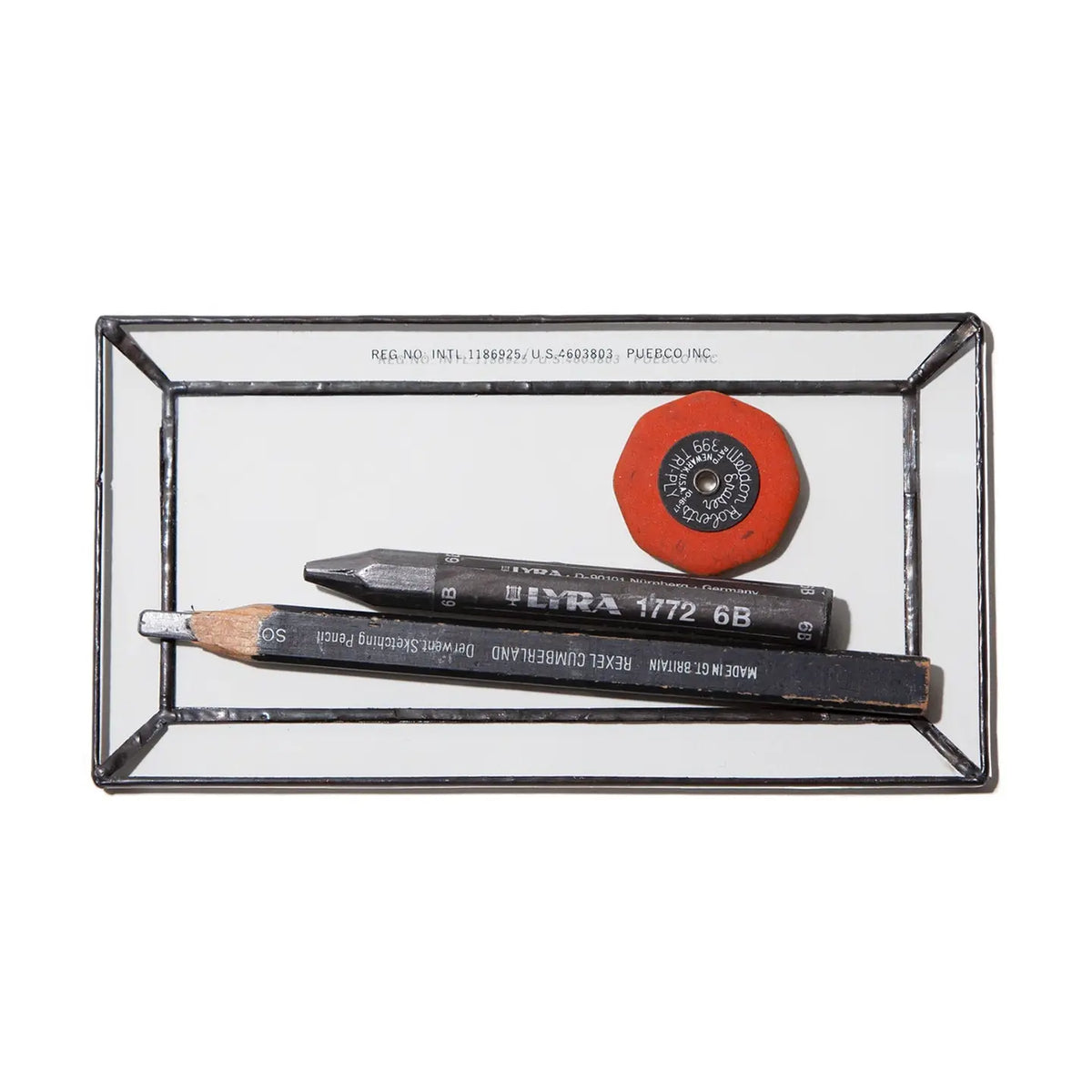 The Faire Glass Valet Tray neatly holds a red eraser, a black woodless pencil, and a sharpened black pencil with white text, offering a sleek and organized storage solution.