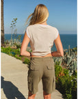 A woman with long blonde hair, wearing the Faire Hotel Luna Muscle Tank and olive green cargo shorts, stands on a sunlit path overlooking the ocean and coastal vegetation.