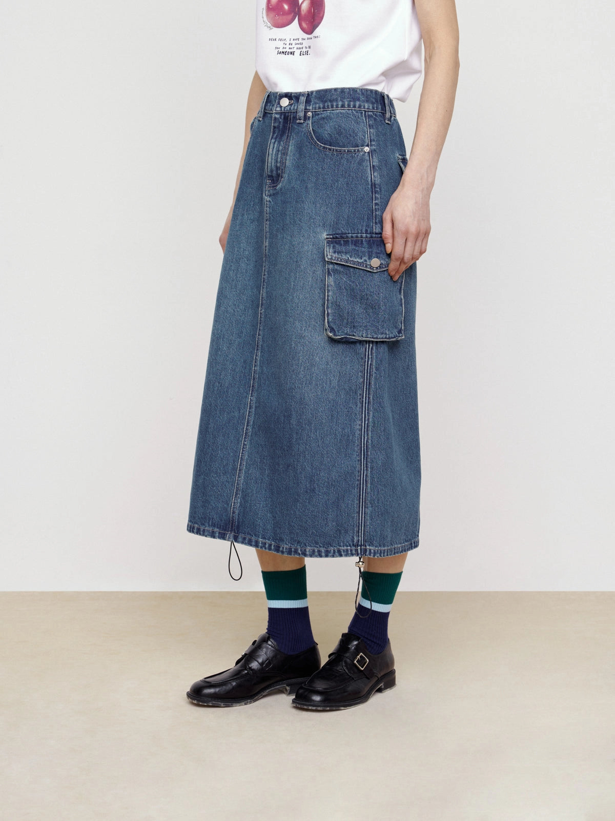 Model wearing Faire’s Pocket Denim Half Skirt styled with a white T-shirt, black shoes, and navy-green striped socks for an effortless street look against a plain backdrop.