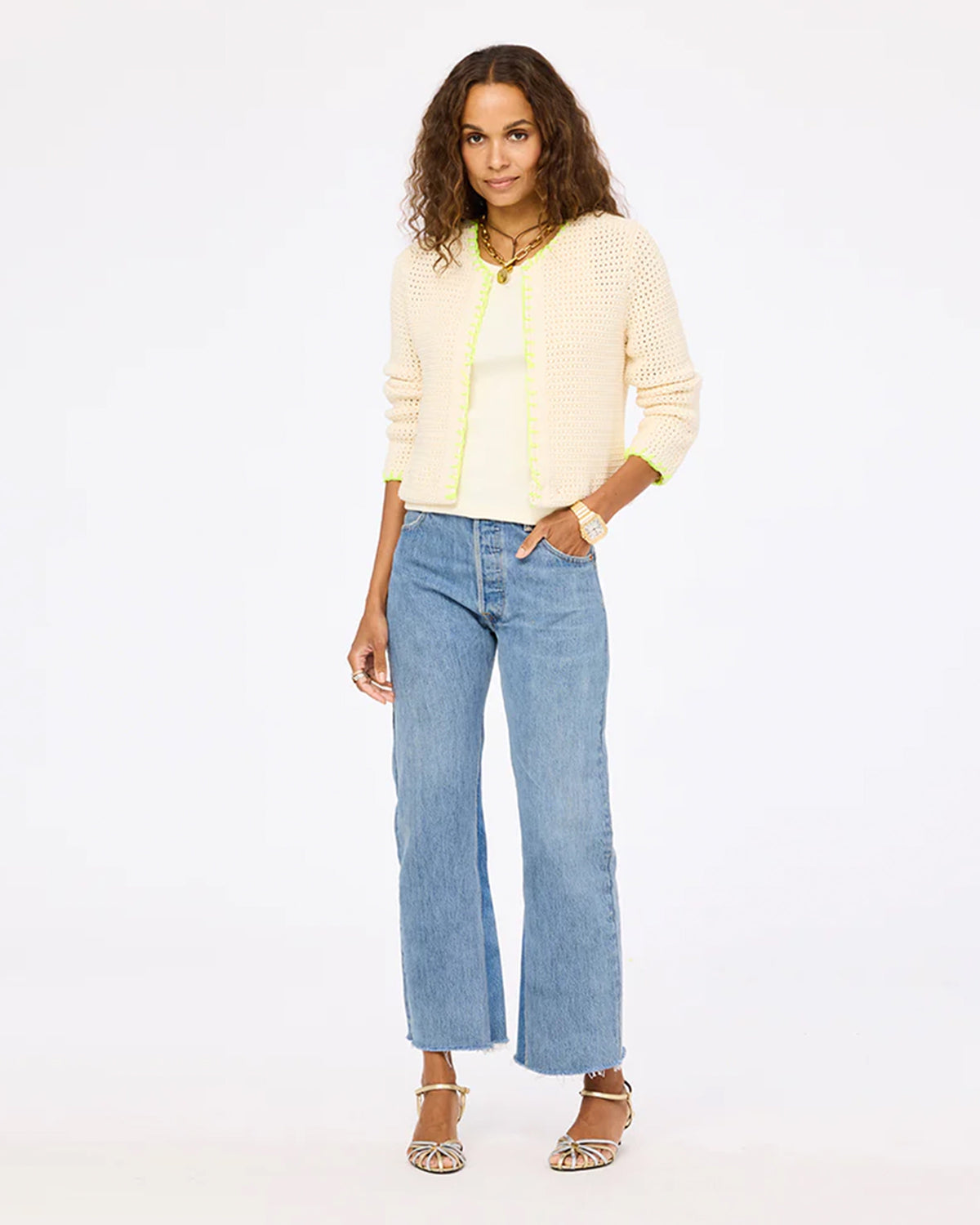 A woman with curly hair poses confidently in the Kerri Rosenthal Frankie Beau-Lero: an open-front cream cardigan with neon trim, styled over a white top, blue wide-leg jeans, strappy sandals, and jewelry against a white backdrop.