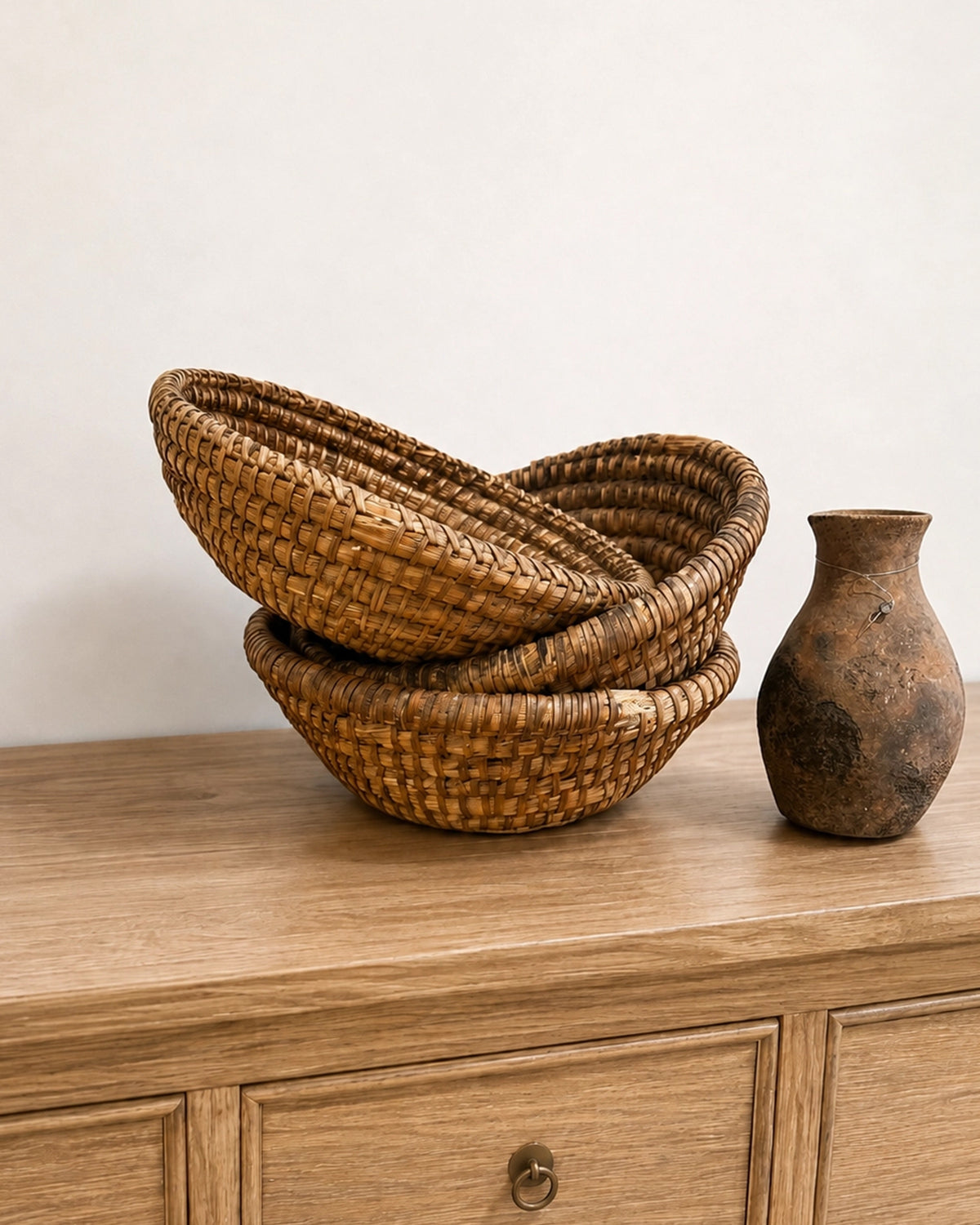 Two stacked vintage French antique baskets and a rustic ceramic vase on a light wood surface from Bungalow Furniture.