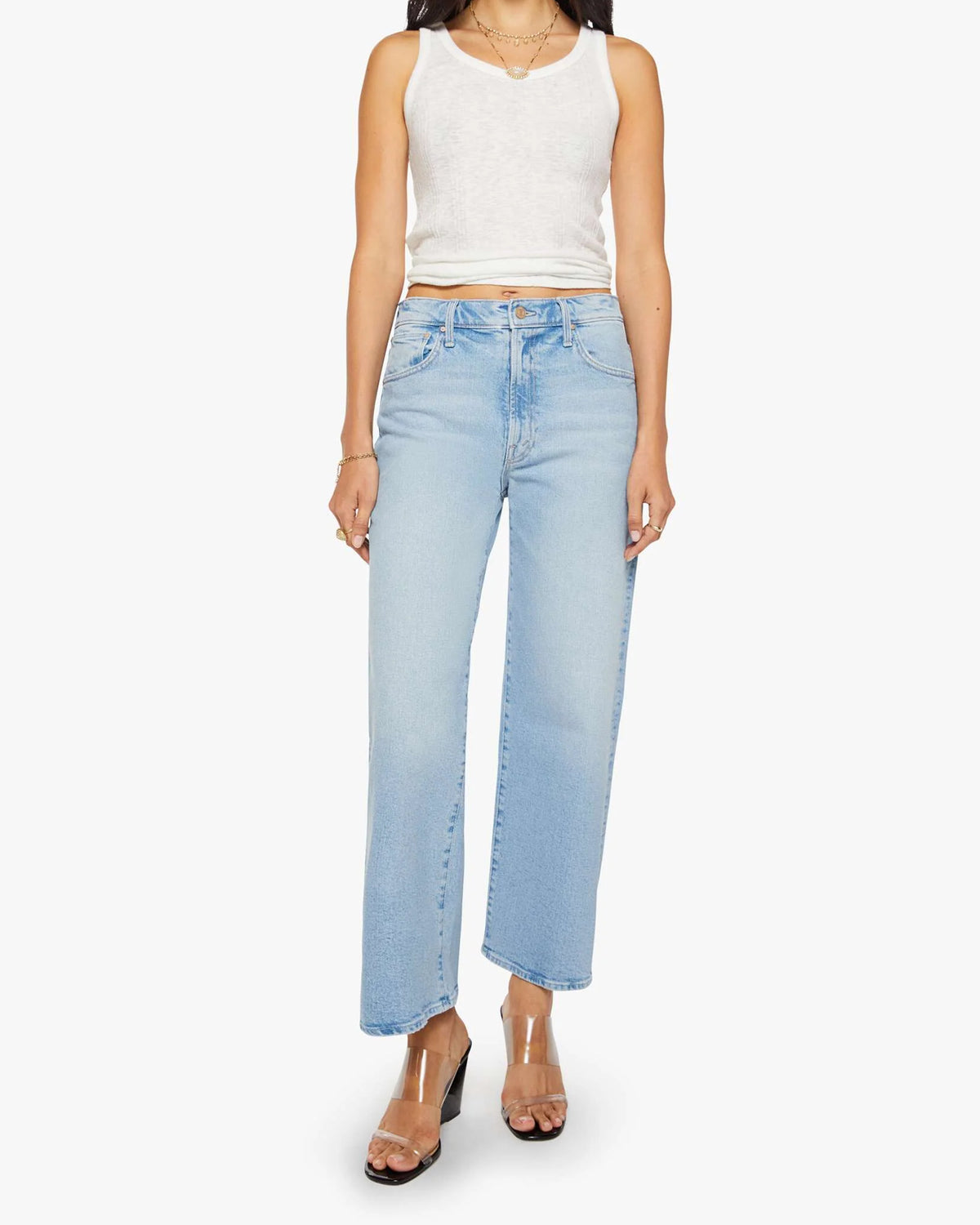 A person in The Dodger Ankle loose straight leg jeans by Mother, paired with a white sleeveless top and clear high-heeled sandals, stands against a white background.