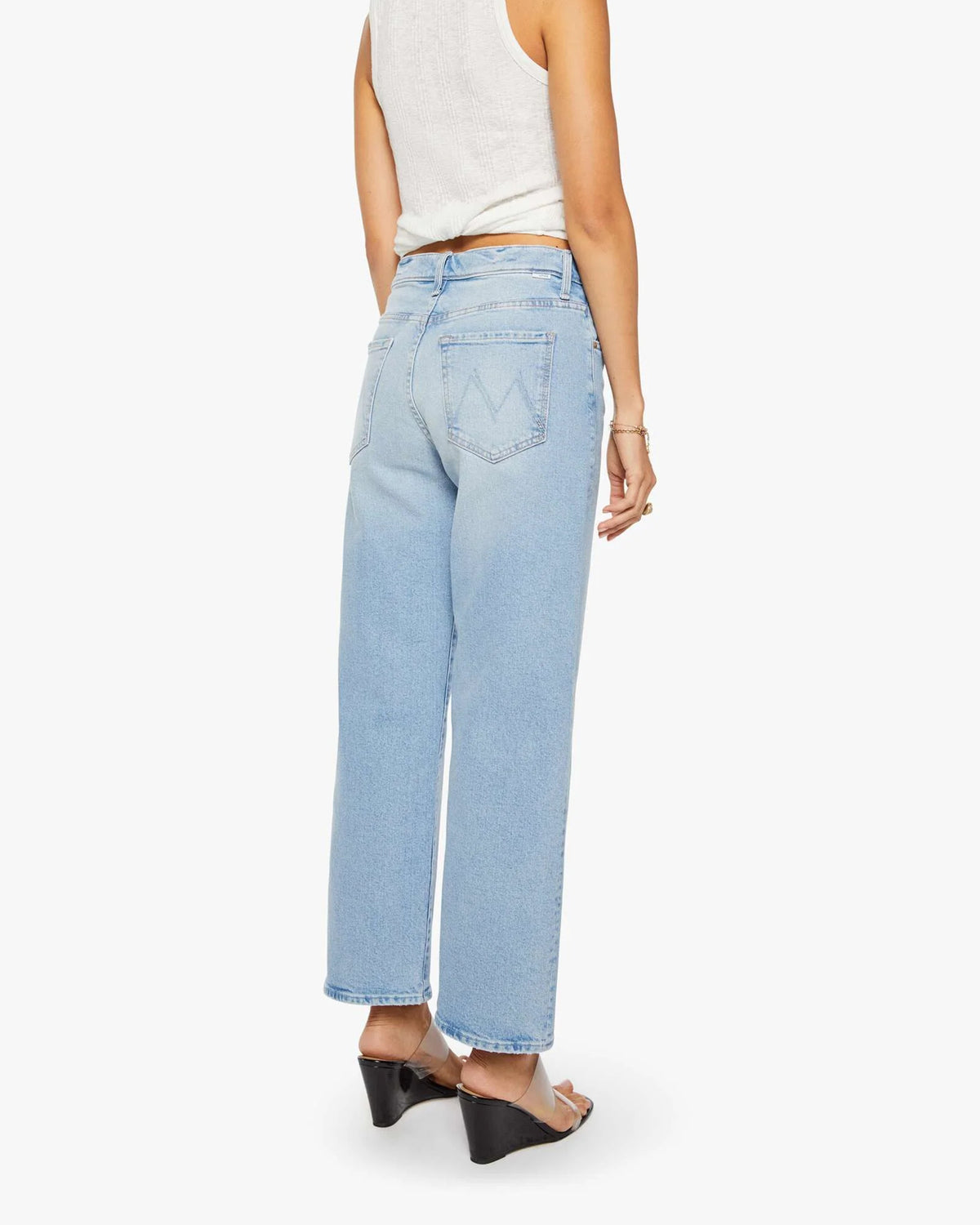 A person stands facing away, showcasing the back of Mother’s The Dodger Ankle high-waisted, ankle-length loose straight leg jeans, paired with a white sleeveless top and black wedge sandals.